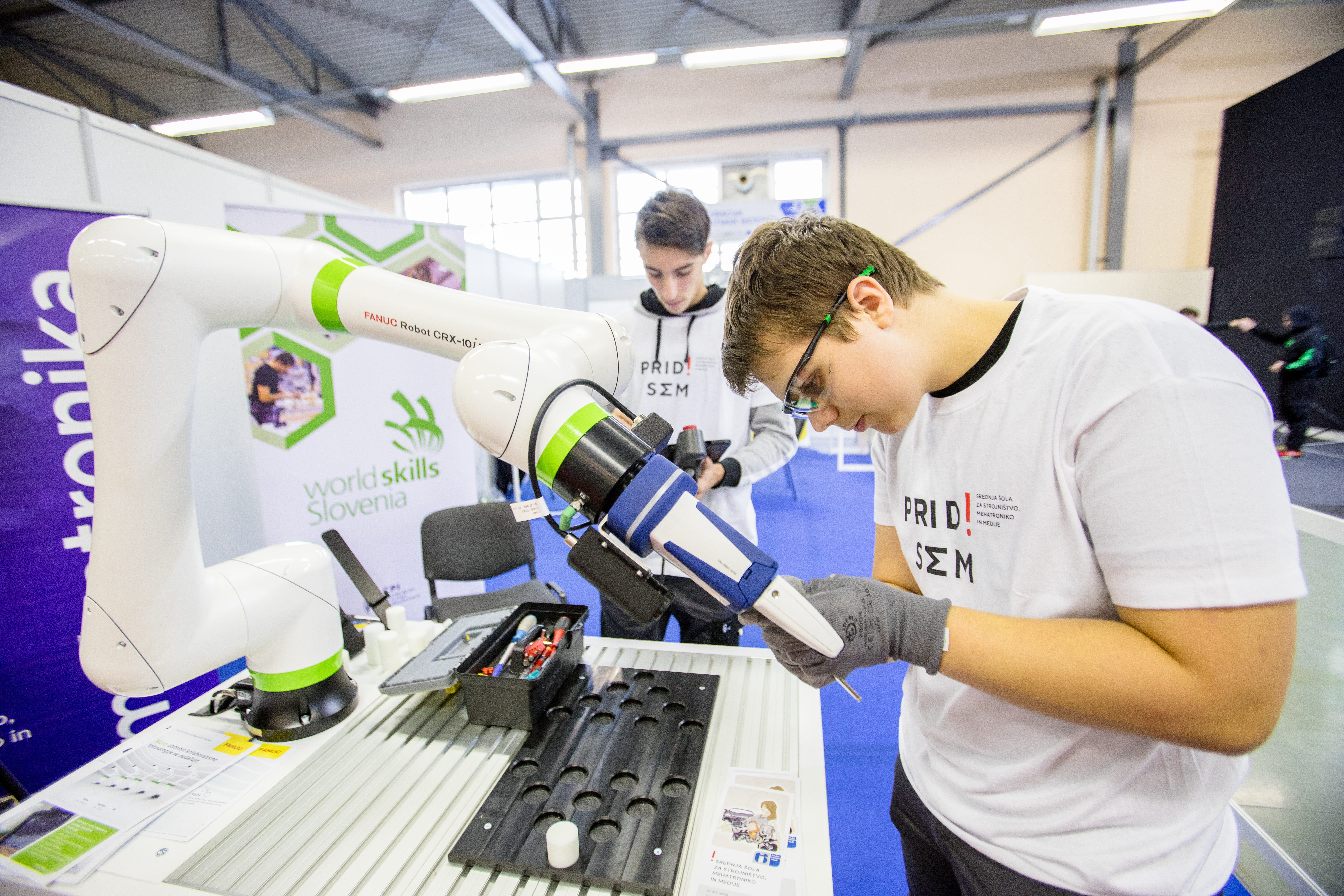 Tekmovalec zateguje prijemala na roki robota. Njgov kolega v ozadju programira robotsko roko na tablici.